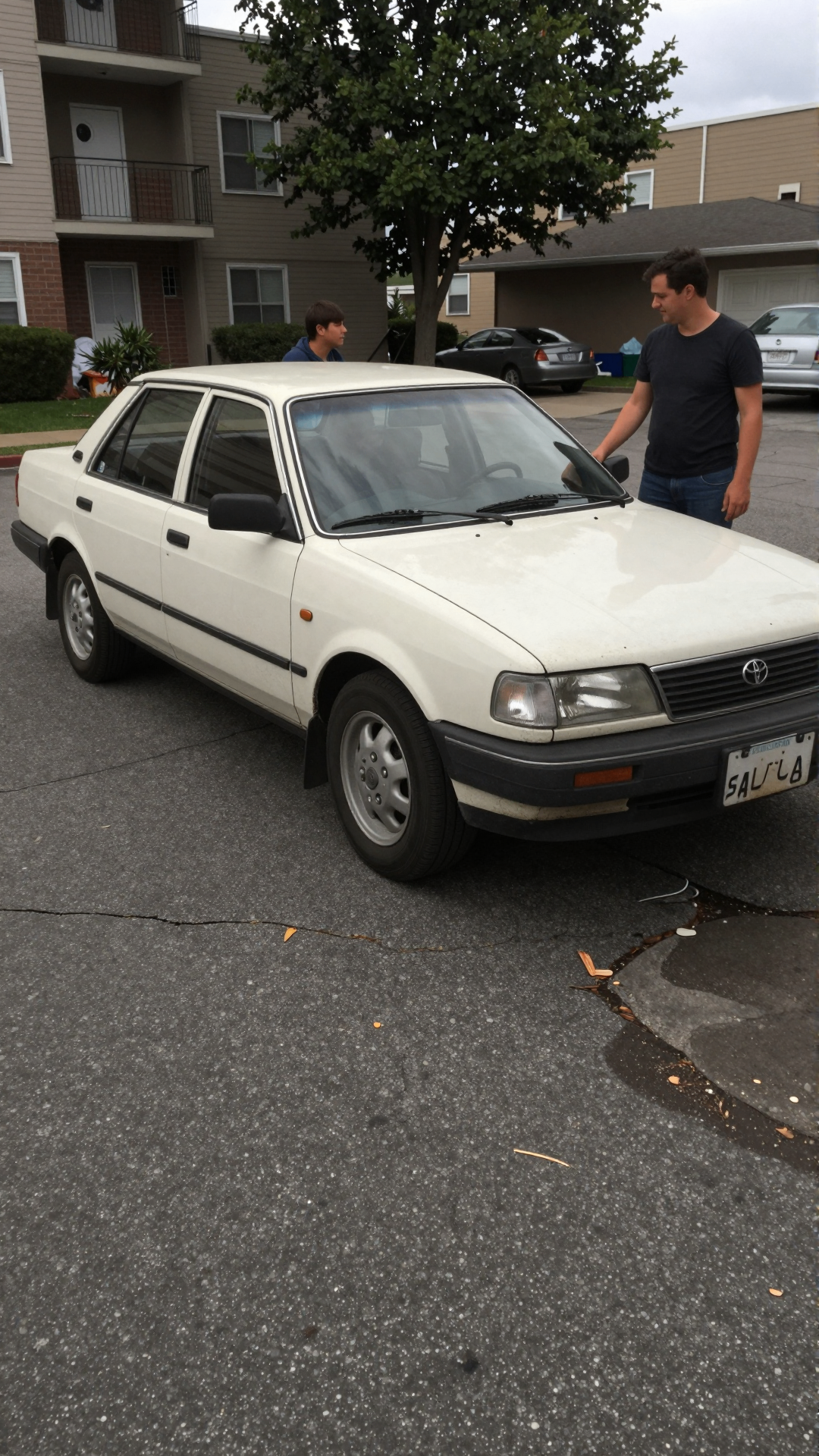 Two men standing beside a white vintage Toyota in an apartment parking lot.